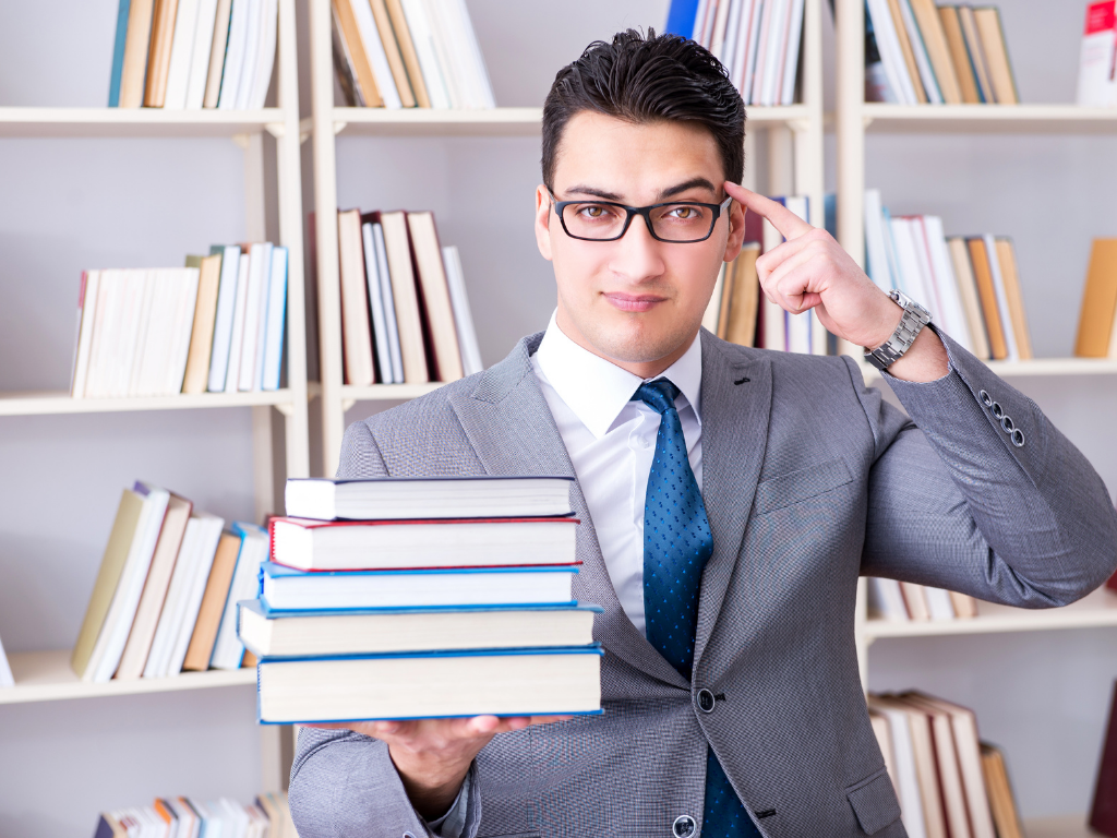 Man in suit with blue tie holding stack of books in one hand and point to temple with other hand