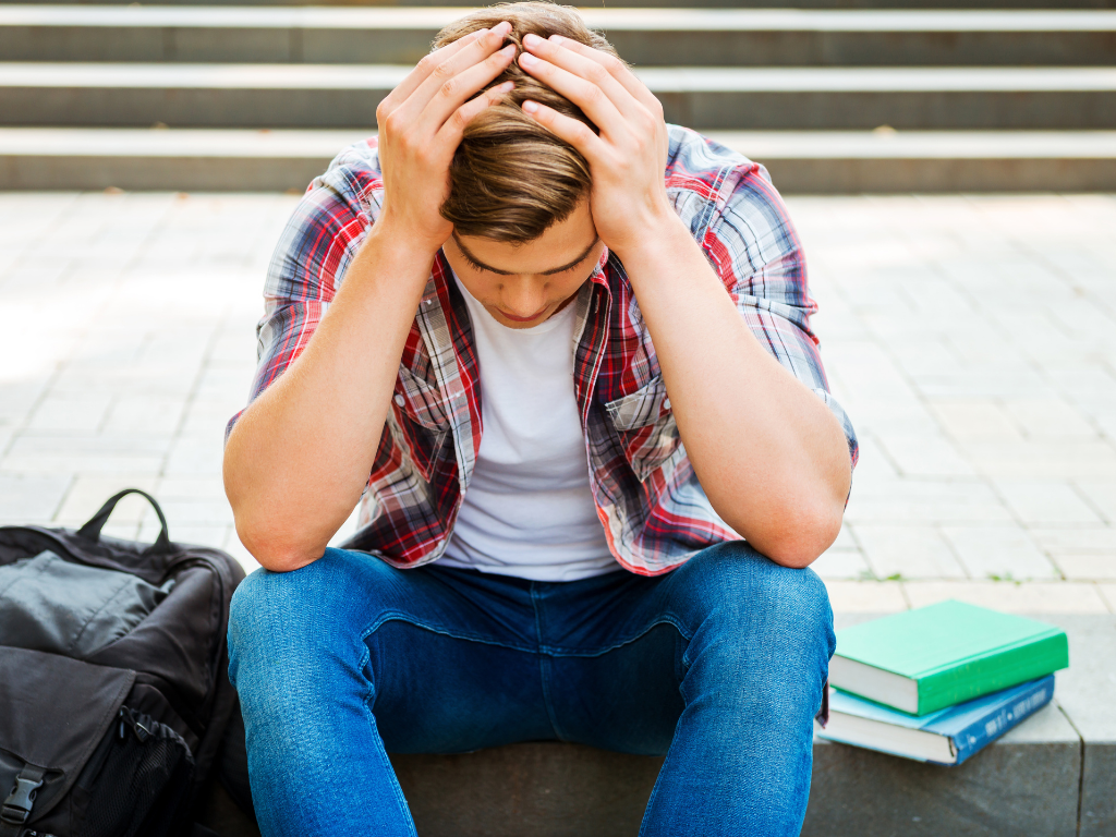 Man sitting on step with head down in hands surrounded by books and a book bag