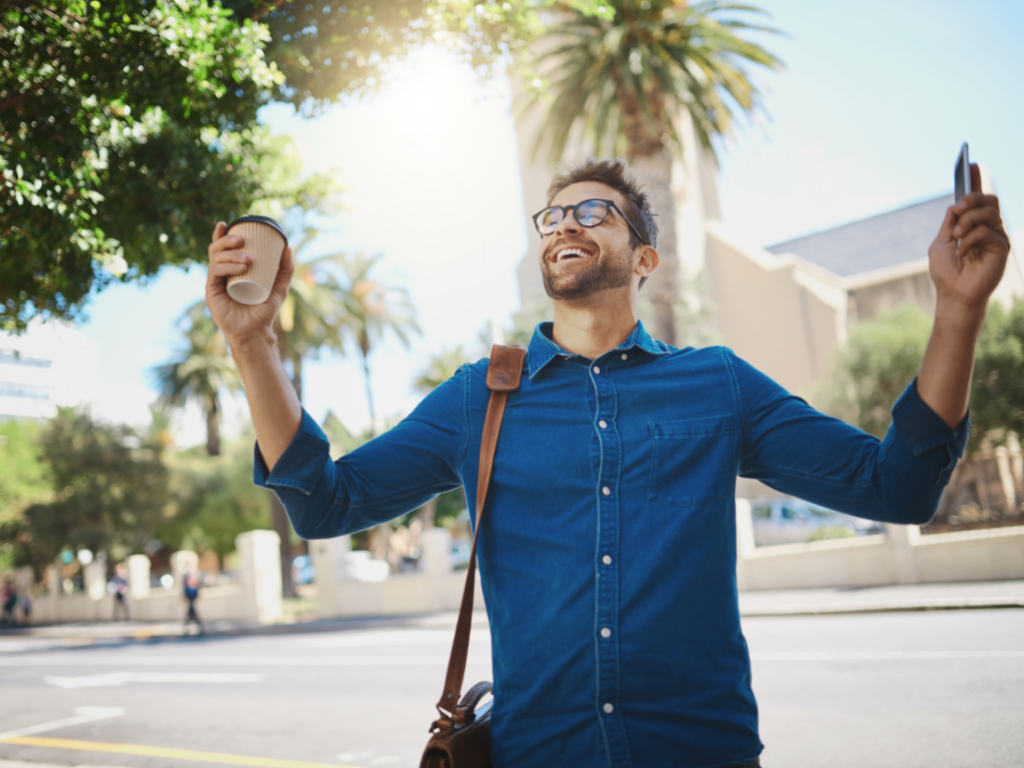 Man in blue shirt and glasses with arms lifted coffee in one hand and cell phone in the other.