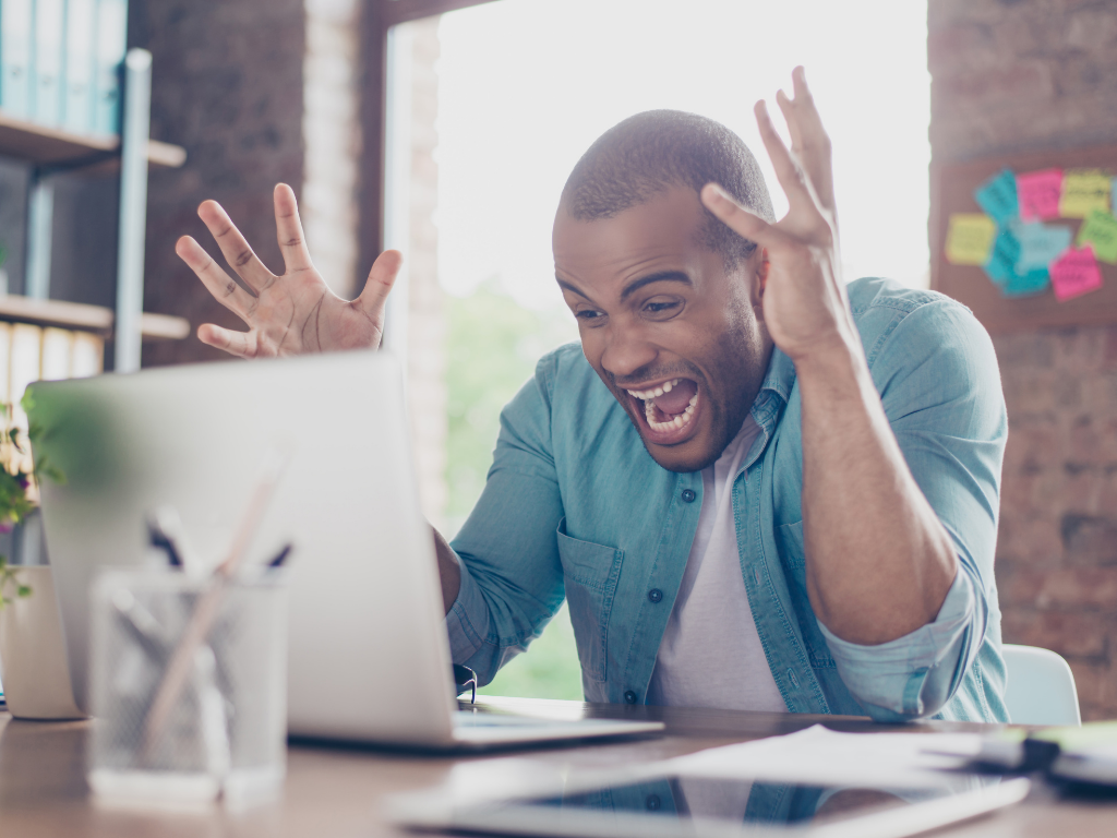 Man yelling at computer in a blue shirt with arms raised and mouth open.