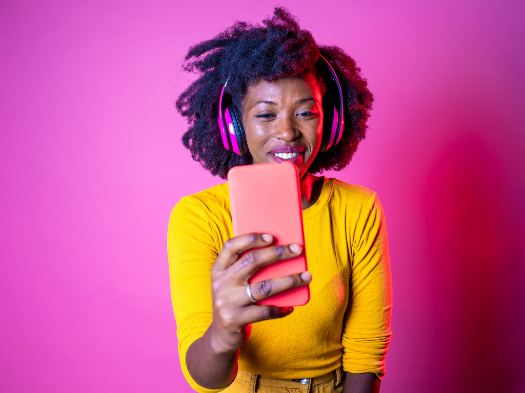  Women looking at phone with headphones on. pink background, and wearing a yellow shirt; bar exam prep