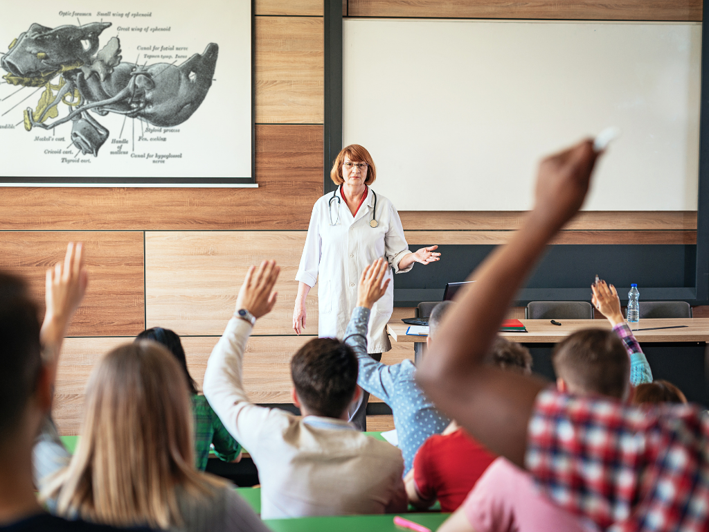 Med school teacher standing at front of class and students have their hands raised in the air. 