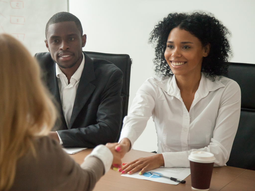 two people shaking hands across a table for a job interview with a 2nd interveiwer overseeing