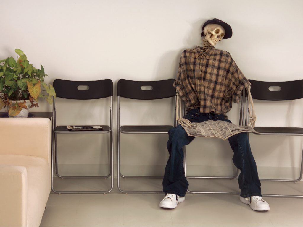 Skeleton in a full outfit sitting in a chair in a waiting room with nespaper in hand