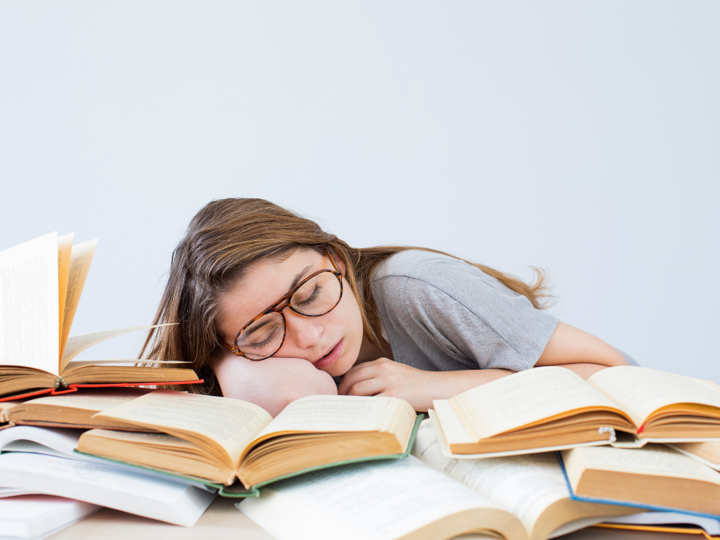 Female with glasses and a grey shirt falling asleep on a pile of books